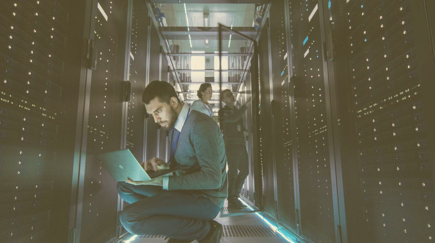 Stock image of a bearded man with a laptop working in a room full of servers.