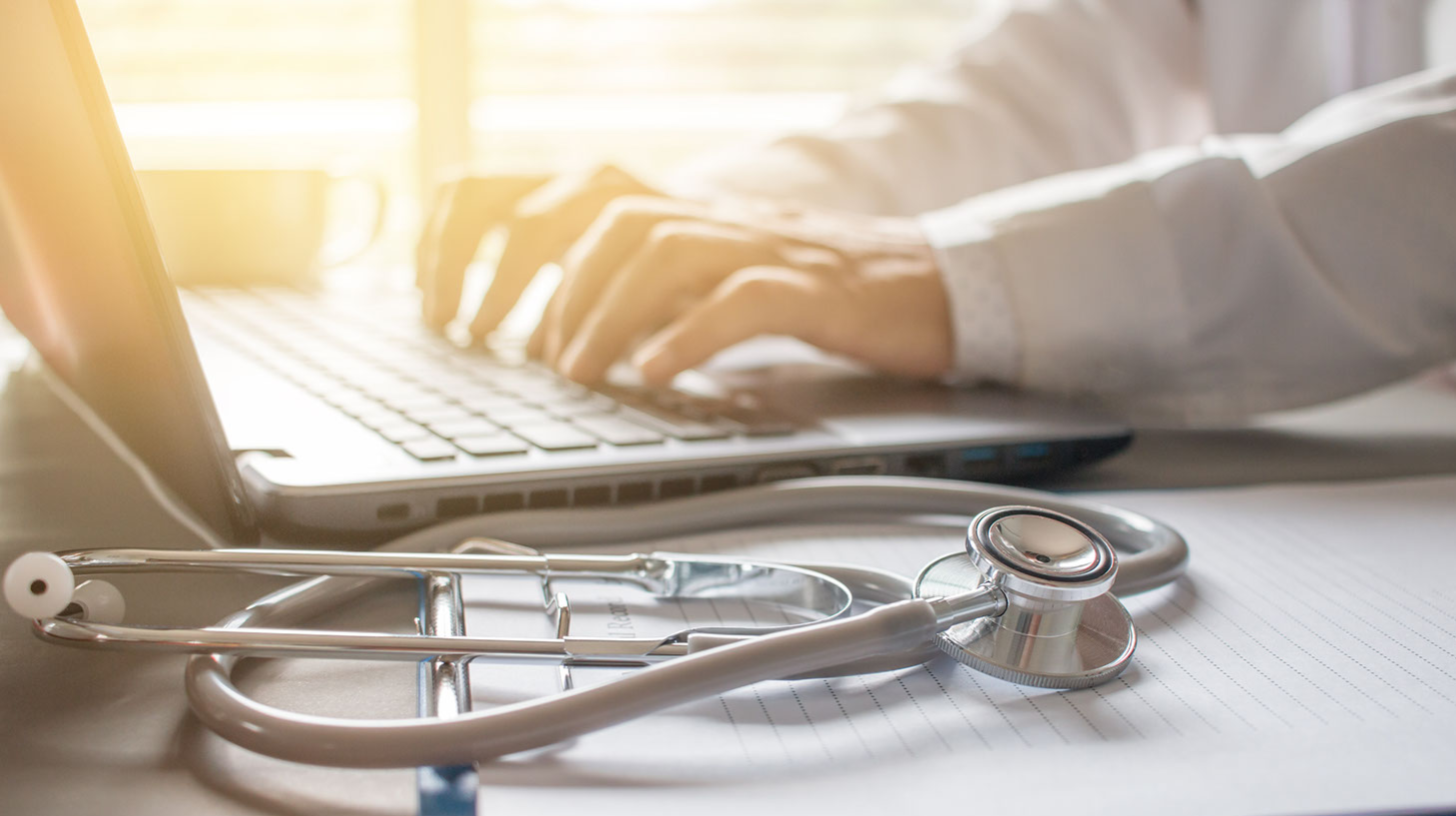 Image of a person in a white doctor's coat typing on a laptop with a stethoscope lying nearby.