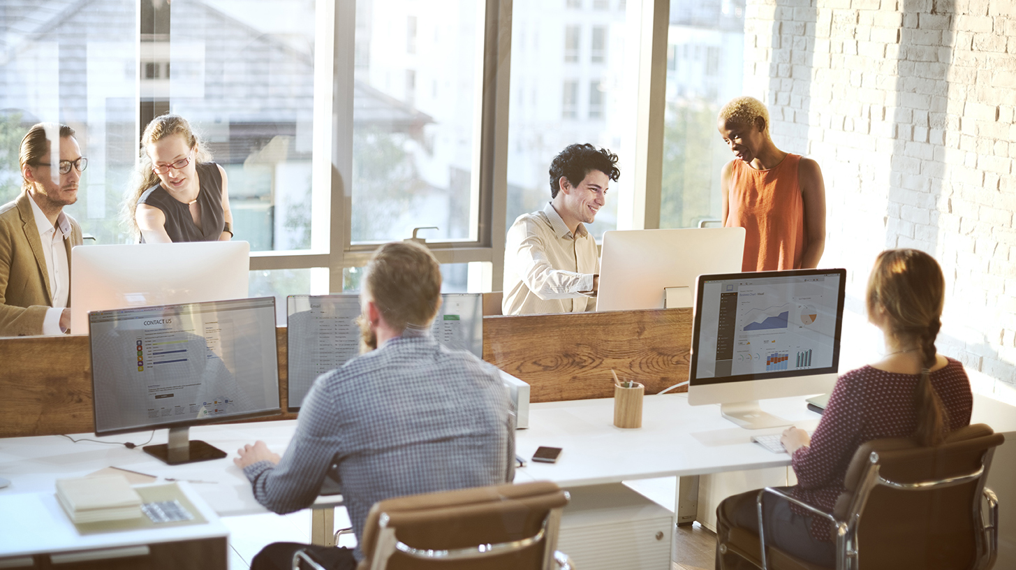 Photo of a group of people working at a conference table with five desktop computers and a large window in the background.