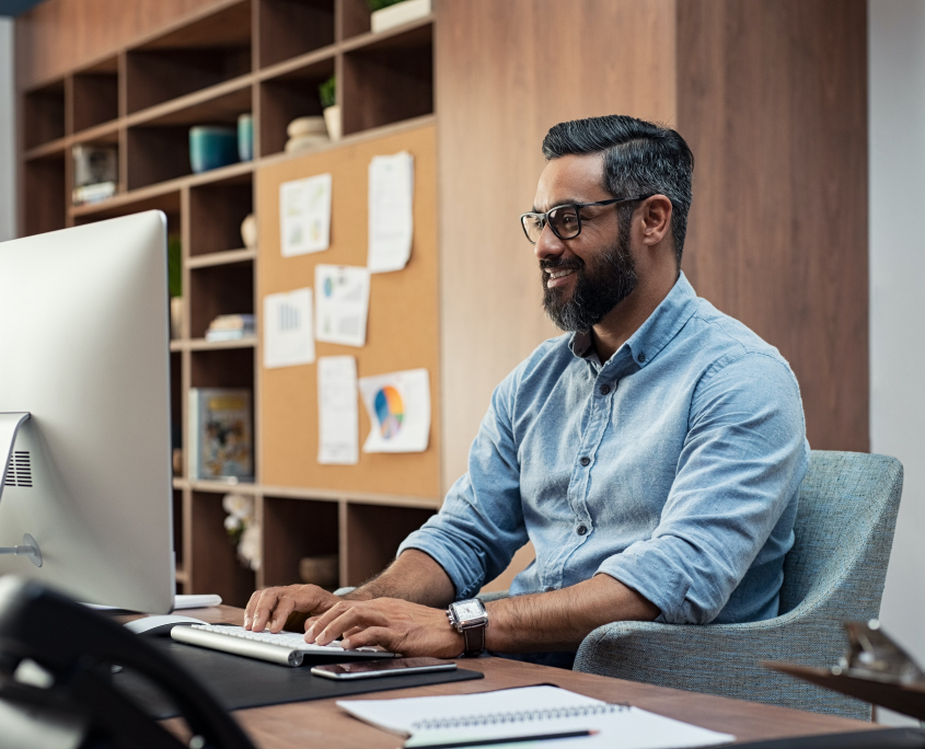 Stock image of a bearded white many in a blue shirt working on a desktop computer.