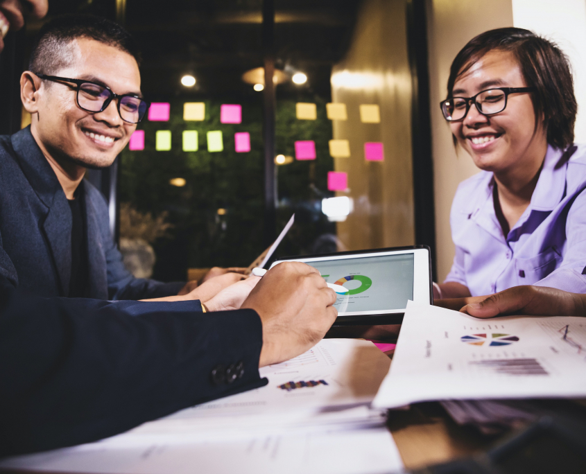 Stock image of an Asian man and an Asian woman smiling and working at a table covered with charts.